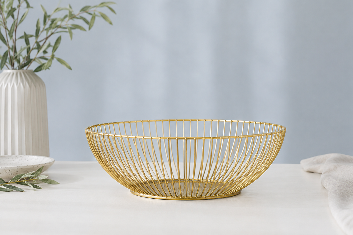 A gold wire bowl with a slatted open design, displayed on a white table with a light blue background, flanked by decorative props including a white vase with greenery.